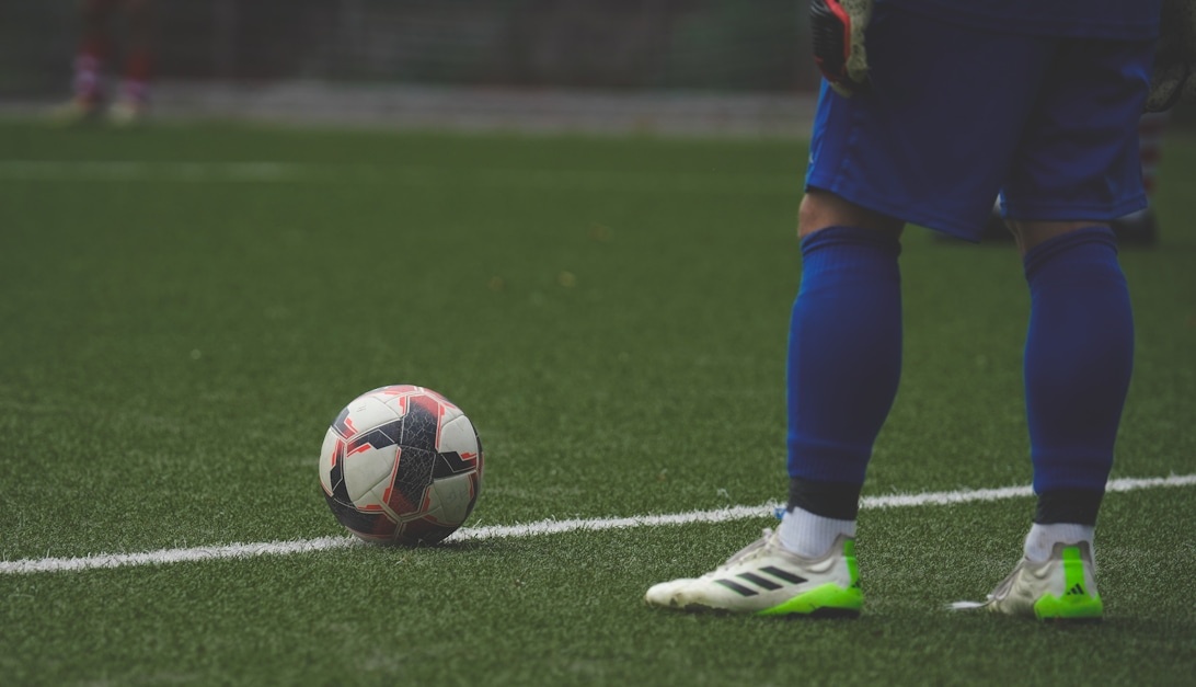 En fotboll på en grön gräsplan med en spelares ben och Adidas-skor synliga, vilket fångar ett aktivt ögonblick under en match.